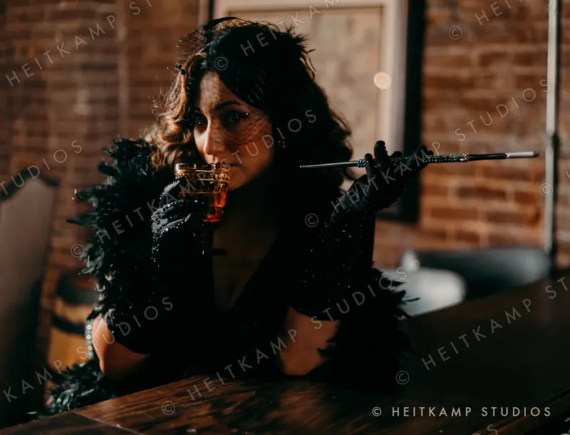 Artemisa sipping a cocktail in a black veil and rhinestone gloves