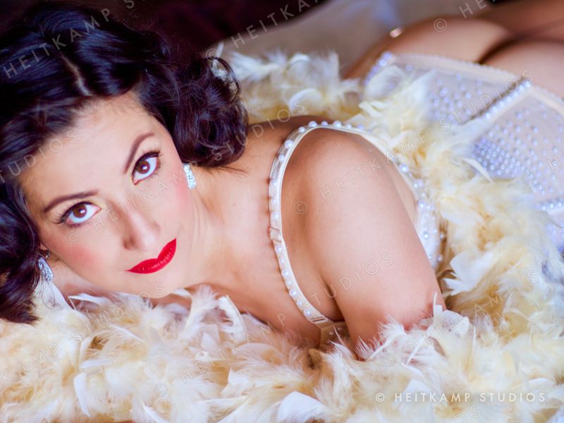 Artemisa lying in cream feathers with red lips and pin curls, looking up at the camera