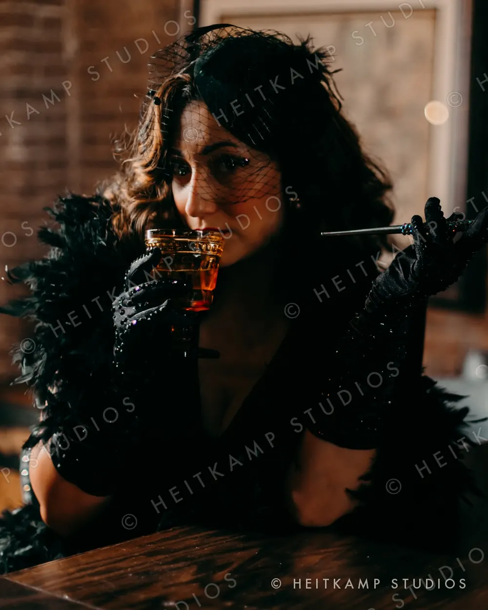 Artemisa in a black veil, feather boa, and rhinestone gloves, sipping a cocktail with a long cigarette holder