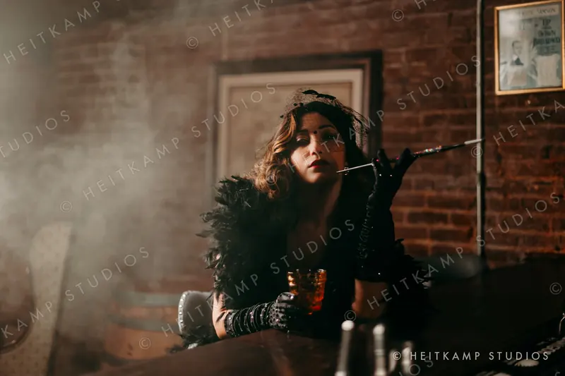 Artemisa in a black veil and feather boa at a smoky bar, holding a jeweled cigarette holder and an amber cocktail