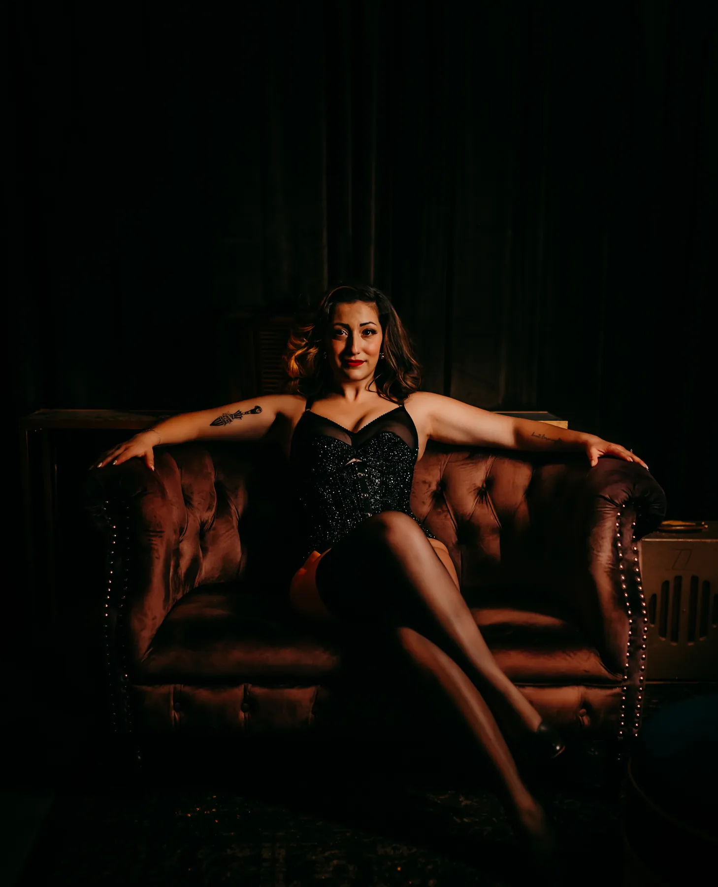 Artemisa lounging on a tufted velvet sofa in a black rhinestone corset and stockings